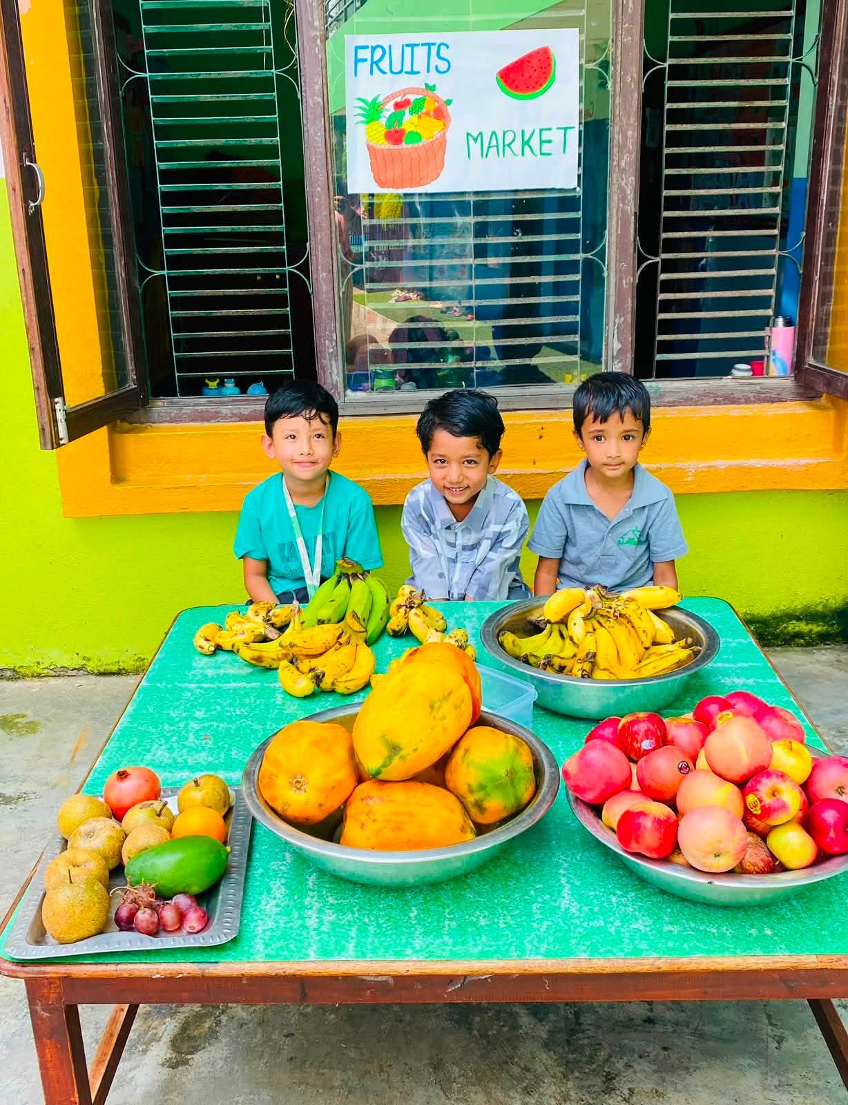 Three students sitting behind their Fruits Market stall of bananas, papayas and apples